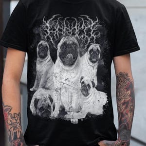 May include: Black t-shirt featuring a graphic of several pugs. The shirt has a distressed, vintage look. The pugs are in various poses, with a stylized font above them. The person wearing the shirt has tattoos and green hair.