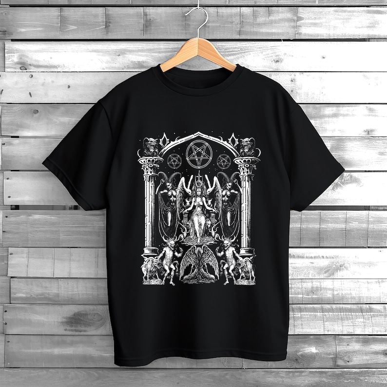 May include: Black t-shirt featuring a detailed white graphic of a gothic scene with a central figure, angels, demons, and a pentagram. The design is intricate and detailed, creating a striking visual on the dark fabric. The shirt is hanging on a wooden hanger.
