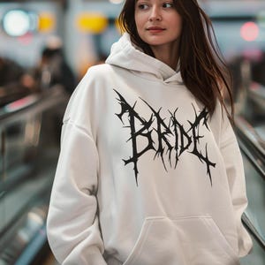 May include: A white hooded sweatshirt featuring a black "BRIDE" graphic in a spiky, gothic font. The sweatshirt has a front pocket and a drawstring hood. The material appears soft.