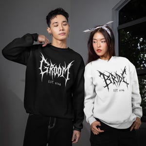 May include: Black and white sweatshirts with gothic lettering. The black sweatshirt reads "GROOM EST. 2024" in white. The white sweatshirt reads "BRIDE EST. 2024" in black. Both have long sleeves.