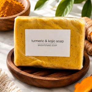 Puede incluir: Una pastilla rectangular de jabón de cúrcuma y ácido kójico envuelta en plástico transparente. La etiqueta blanca en la parte frontal presenta el texto negro "turmeric & kojic soap" y "BRIGHTENING SOAP" en una fuente sans-serif clara.