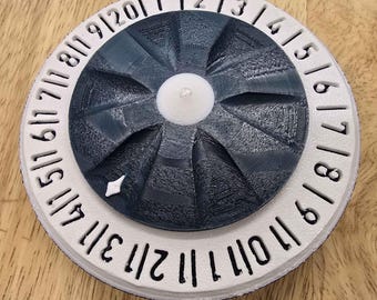 Puede incluir: Una rueda de calendario impresa en 3D, blanca y gris oscuro, con los números del 1 al 12 en el borde. El centro tiene un botón blanco y un diseño en forma de abanico gris oscuro. Los números son en negro.