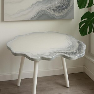 May include: A small, white side table with a unique, cloud-like shape. The tabletop features a marbled design in shades of gray and white, with a silver-colored edge. The table stands on three white legs.