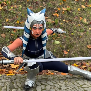 May include: A person in a Star Wars Ahsoka Tano costume, kneeling on a stone surface. The costume includes a blue and white headpiece, face paint, and a navy blue outfit with silver accents. Two lightsabers are held.