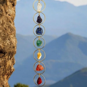 May include: A chakra stone wind chime featuring seven colorful raw crystals suspended within gold-colored hoops. The crystals include clear, dark blue, light blue, green, yellow, orange, and red stones. The wind chime hangs from a brown cord against a backdrop of mountains.