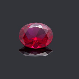 May include: A deep red oval-shaped gemstone with a faceted cut. The gemstone is set against a black background.