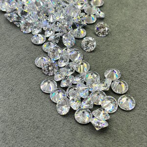 May include: A large quantity of clear, faceted, round gemstones, likely diamonds, are scattered on a gray surface. Some of the gemstones are in a clear plastic bag.