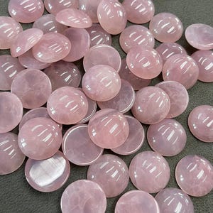 May include: A collection of polished rose quartz stones in various shapes. The stones are primarily round and flat, with a soft pink hue and a glossy finish. Some stones are domed, while others are flat.