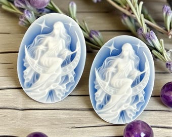 May include: Two oval-shaped, blue and white cameo pendants. Each pendant features a carved image of a woman seated on a crescent moon, surrounded by stars. The pendants are displayed on a wooden surface, with purple gemstones and lavender sprigs.