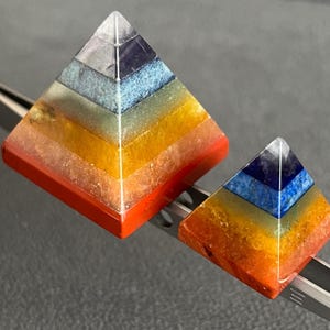 May include: Two multi-colored crystal pyramids, one larger than the other, are arranged on a silver tool. The pyramids are made of seven layers of different colored crystals, including red, orange, yellow, green, blue, indigo, and violet.