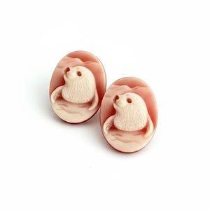 May include: A pair of oval-shaped cameo earrings featuring a carved cream-colored seal motif set against a smooth, dusty rose-colored background. The seals are depicted in profile, facing upward, with textured fur details and rounded flippers.