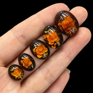 May include: Five oval-shaped amber cabochons with pressed orange flowers inside. The cabochons are arranged in a row from largest to smallest.