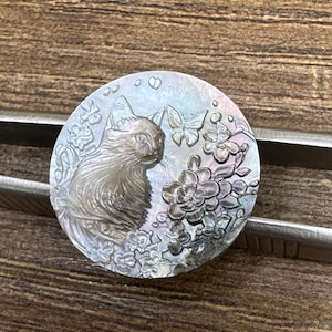 May include: A round, iridescent cat-themed pendant. The pendant features a detailed relief of a cat, butterflies, and flowers. The design is carved into a mother-of-pearl-like material, creating a shimmering effect. The pendant is held by a pair of metal tweezers.
