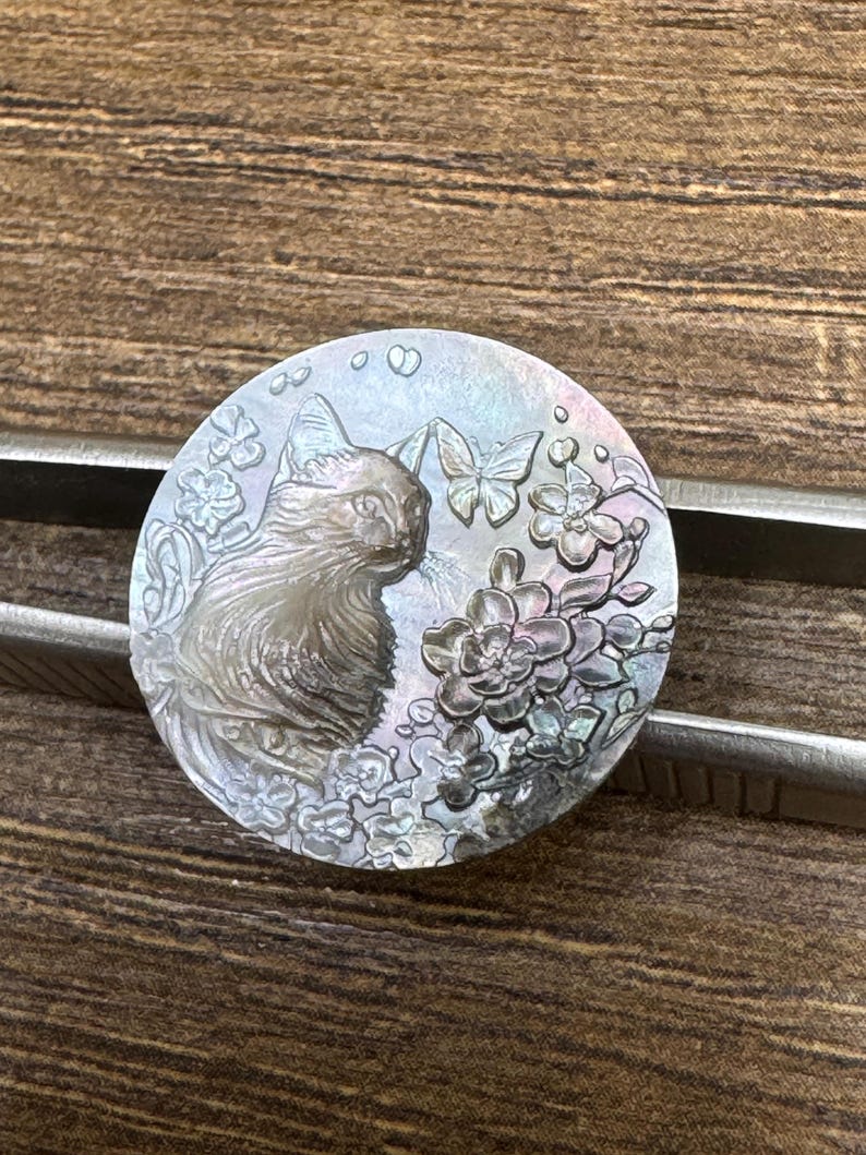 May include: A round, iridescent cat-themed pendant. The pendant features a detailed relief of a cat, butterflies, and flowers. The design is carved into a mother-of-pearl-like material, creating a shimmering effect. The pendant is held by a pair of metal tweezers.