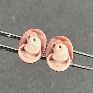 May include: A pair of oval-shaped cameos featuring a carved relief of a seal in a light cream color against a soft pink background. The two identical pieces are held by a pair of silver-toned metal tweezers, showcasing the detailed animal motif.