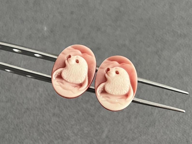 May include: A pair of oval-shaped cameos featuring a carved relief of a seal in a light cream color against a soft pink background. The two identical pieces are held by a pair of silver-toned metal tweezers, showcasing the detailed animal motif.
