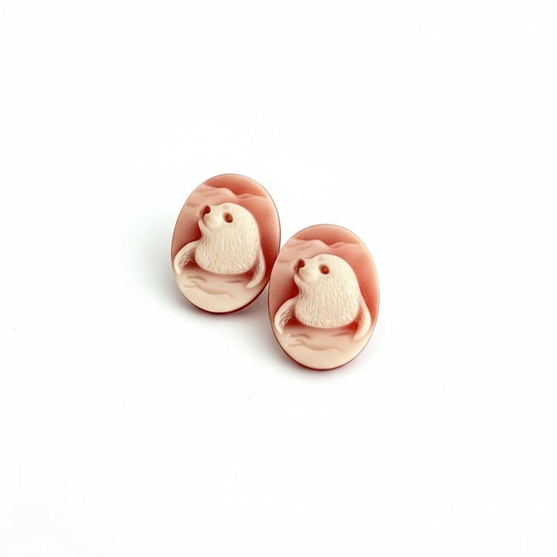 May include: A pair of oval-shaped cameo earrings featuring a carved cream-colored seal motif set against a smooth, dusty rose-colored background. The seals are depicted in profile, facing upward, with textured fur details and rounded flippers.