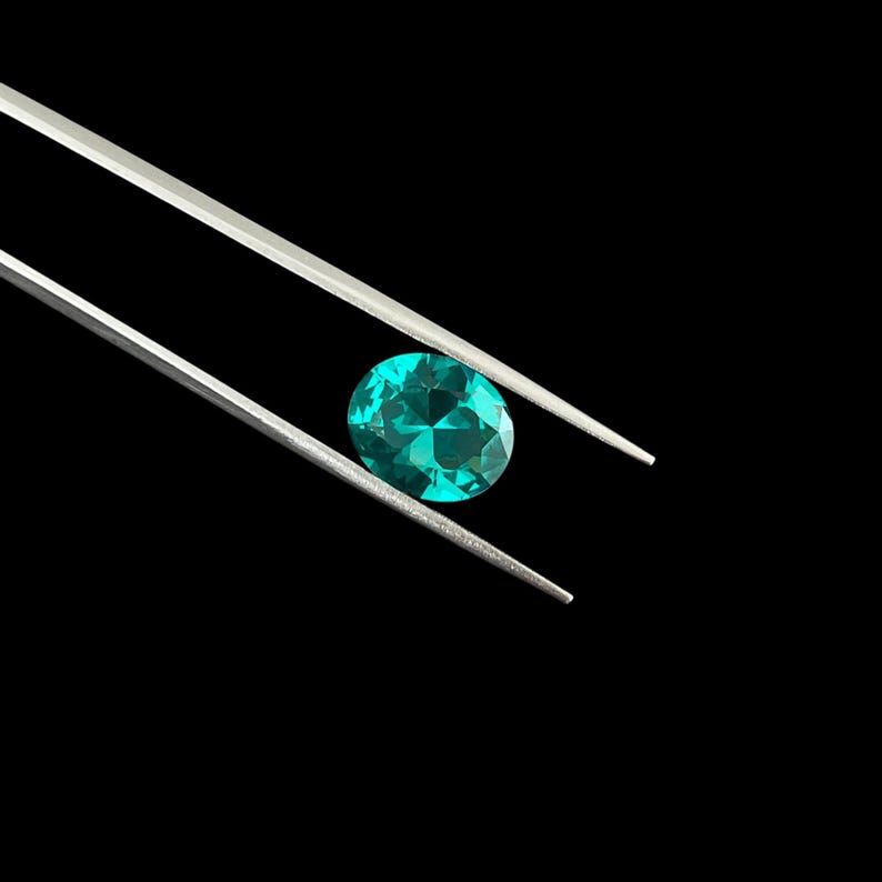 May include: A vibrant, oval-cut teal gemstone held securely by a pair of silver-toned metal tweezers. The gemstone features a faceted surface that reflects light, displaying a deep, saturated blue-green color against a solid black background.