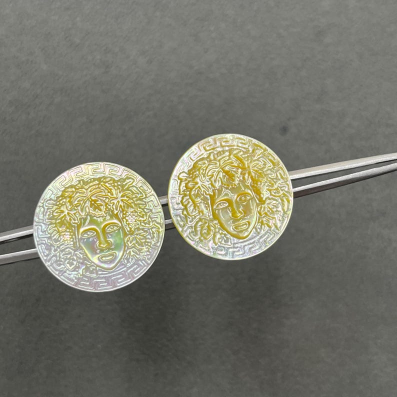 May include: Two circular mother-of-pearl earrings featuring an iridescent finish and a central relief of a face surrounded by leaves. Each piece is framed by a geometric Greek key border pattern and is held by silver-toned metal tweezers.