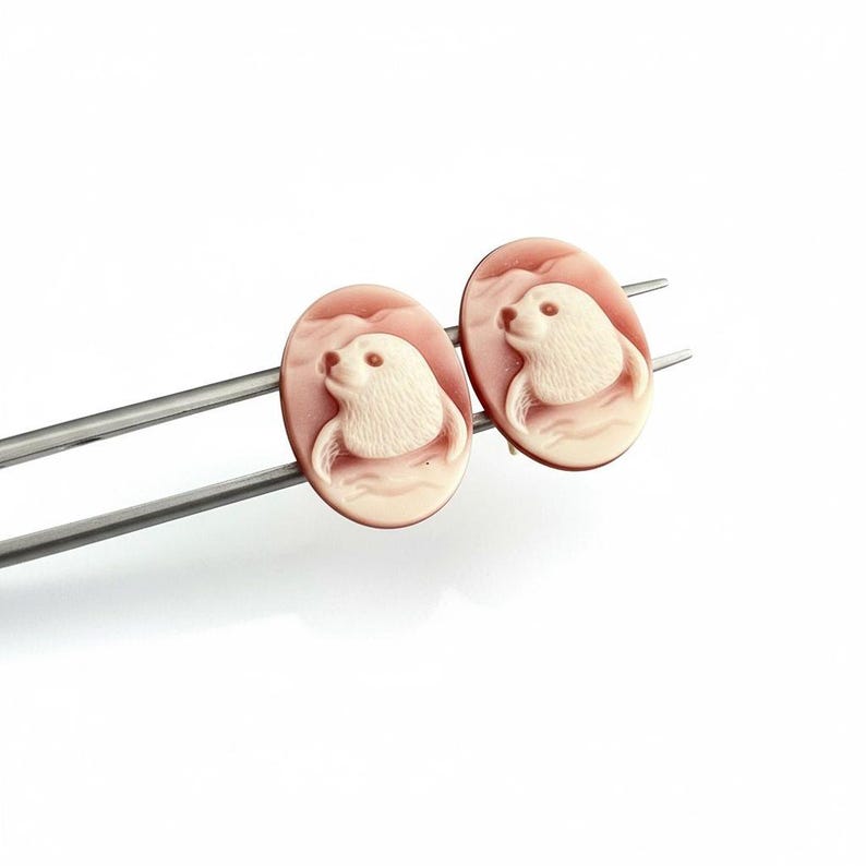 May include: A pair of oval-shaped cameo earrings featuring a carved white seal motif set against a soft pink background. The earrings are mounted on silver-toned metal posts, showcasing detailed relief work of the animal's head and flippers.