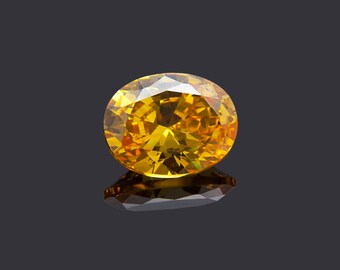 May include: A faceted, oval-cut gemstone with a brilliant yellow hue. The gem is set against a dark gray background, with a reflective surface below. The gemstone's facets catch the light, creating a sparkling effect.