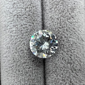 May include: A clear round diamond gemstone with a brilliant cut, set against a gray fuzzy background.