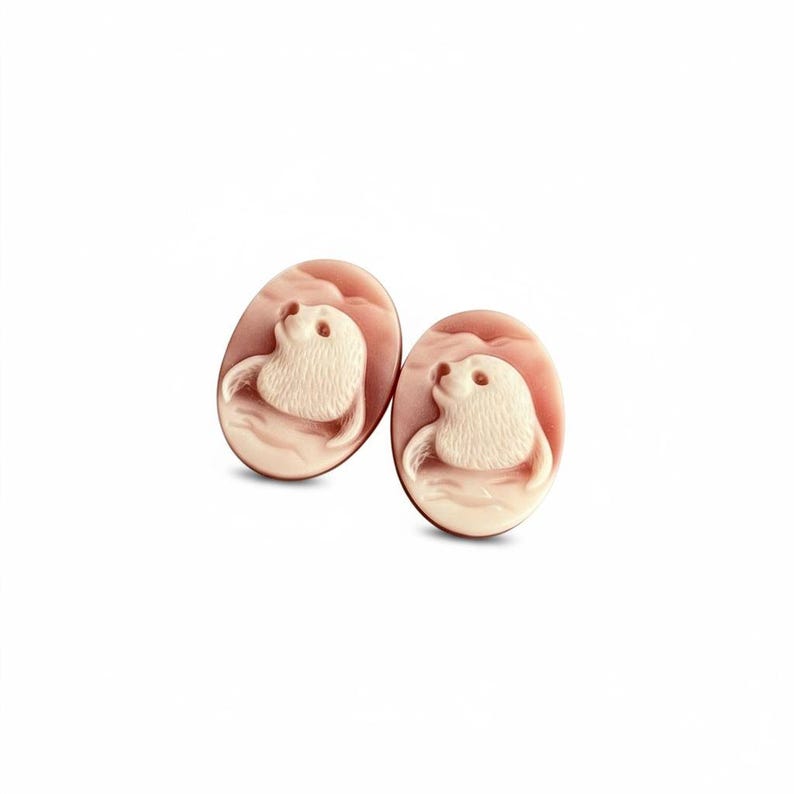 May include: Two oval-shaped cameos featuring a carved cream-colored seal motif set against a soft pink background. Each piece displays a detailed relief of a seal head and flipper, showcasing smooth textures and distinct layered color contrast.