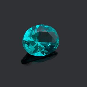 May include: A faceted oval gemstone in a vibrant teal blue color. The stone features a brilliant cut with multiple geometric surfaces that reflect light, creating a sparkling appearance with deep and light blue tones throughout the crystal structure.