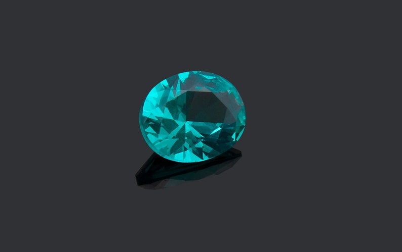 May include: A faceted oval gemstone in a vibrant teal blue color. The stone features a brilliant cut with multiple geometric surfaces that reflect light, creating a sparkling appearance with deep and light blue tones throughout the crystal structure.