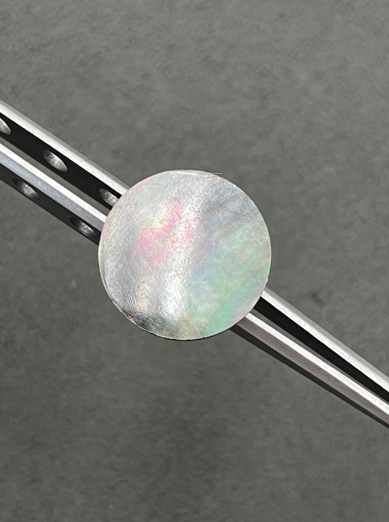 May include: A round, iridescent, mother-of-pearl disc, approximately 1 inch in diameter, is held by a pair of silver metal tweezers. The disc reflects shades of pink, green, and silver. The background is a solid gray.