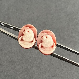 May include: Two oval-shaped cameos featuring a carved light-colored seal motif set against a smooth, coral-pink background. The cameos are held by a pair of silver-toned metal tweezers, showcasing the detailed relief of the marine animal.