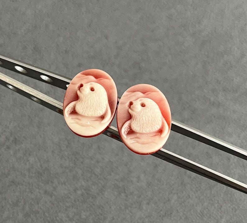 May include: Two oval-shaped cameos featuring a carved light-colored seal motif set against a smooth, coral-pink background. The cameos are held by a pair of silver-toned metal tweezers, showcasing the detailed relief of the marine animal.