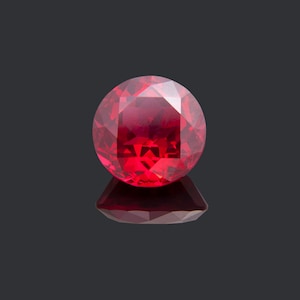 May include: A deep red gemstone with a round cut and many facets. The gemstone is set against a black background.