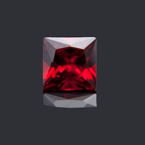 May include: A deep red square-shaped gemstone with a faceted cut. The gemstone is reflecting light on a black background.