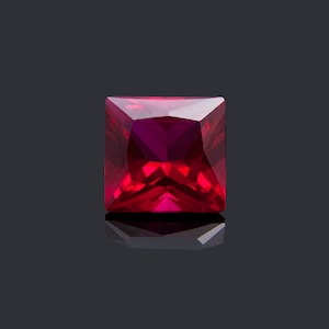 May include: A deep red square-shaped gemstone with a faceted cut. The gemstone is reflecting light and has a shiny finish.
