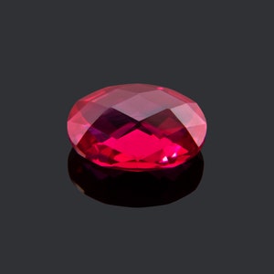 May include: A deep red oval-shaped gemstone with a faceted cut. The gemstone is reflecting light on a black surface.