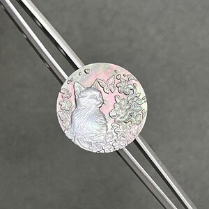 May include: A round, iridescent pendant featuring a detailed cat illustration surrounded by flowers and butterflies. The pendant has a pearlescent sheen with pink and white hues. The pendant is attached to a silver-colored metal object.