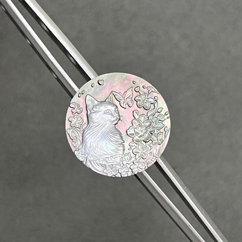 May include: A round, iridescent pendant featuring a detailed cat illustration surrounded by flowers and butterflies. The pendant has a pearlescent sheen with pink and white hues. The pendant is attached to a silver-colored metal object.