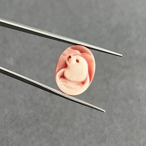 May include: An oval-shaped, light pink cameo carving featuring a relief of a seal. The piece is held by a pair of silver-toned metal tweezers, showcasing the detailed texture of the animal's fur and its rounded form against a smooth background.