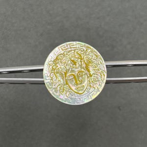 May include: A circular mother-of-pearl button featuring an iridescent finish and a carved relief of a face surrounded by foliage. The outer edge of the button is decorated with a classic Greek key pattern, held by silver-toned metal tweezers.