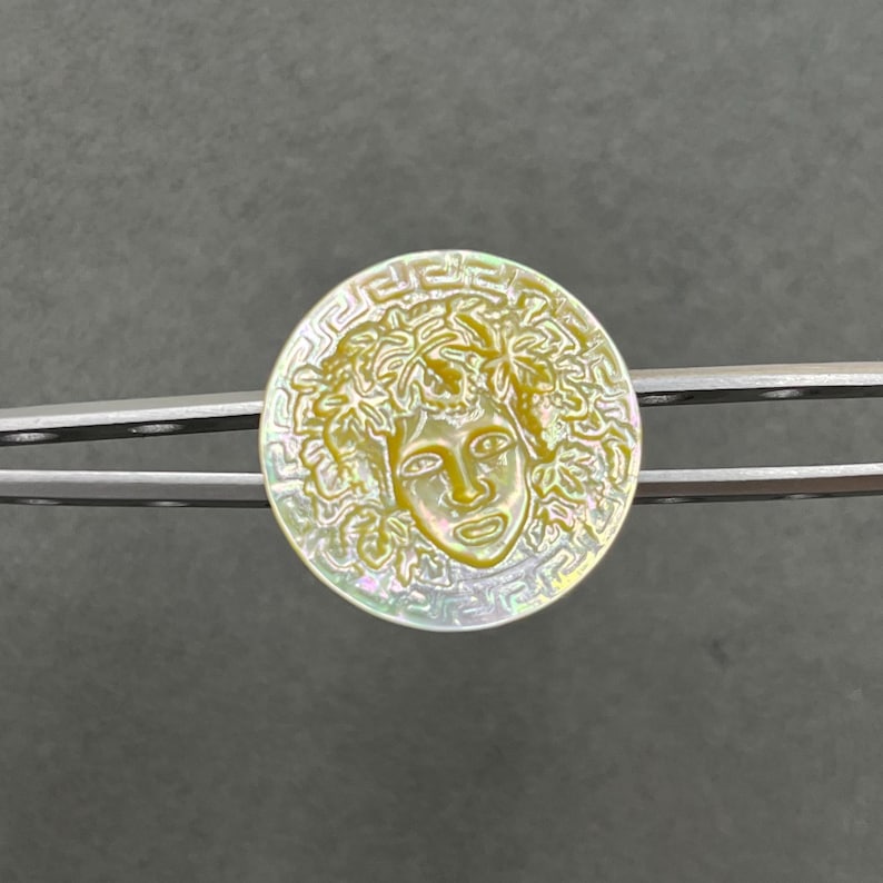 May include: A circular mother-of-pearl button featuring an iridescent finish and a carved relief of a face surrounded by foliage. The outer edge of the button is decorated with a classic Greek key pattern, held by silver-toned metal tweezers.