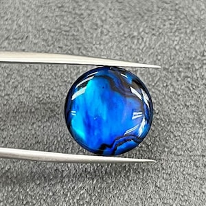 May include: A round, iridescent blue abalone shell cabochon with black and green swirls. The shell is smooth and polished.