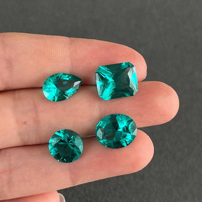 May include: Four faceted loose gemstones in a vibrant teal green color. The collection includes one pear-shaped stone, one rectangular cushion-cut stone, one round brilliant-cut stone, and one oval-shaped stone, all displaying clear internal light refraction.