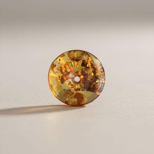 Puede incluir: Una cuenta redonda de color ámbar con un agujero central. La cuenta translúcida revela inclusiones internas en tonos marrones y dorados. La superficie de la cuenta es lisa y pulida, reflejando la luz.