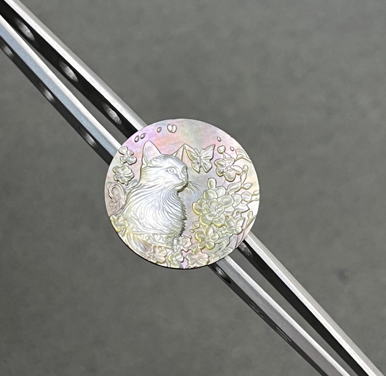 May include: A round, iridescent cat pendant with a floral and butterfly design. The cat is in profile, surrounded by flowers and butterflies. The pendant is set on a silver-colored metal bar.
