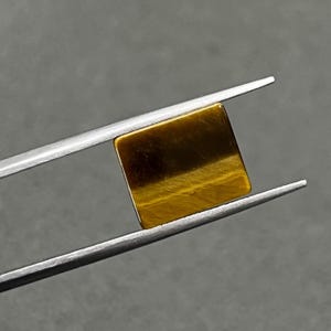 May include: A square-shaped, golden brown tiger's eye gemstone with a chatoyant effect. The gemstone is held in a pair of tweezers.