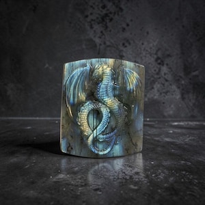 May include: A rectangular, carved stone with a detailed dragon design. The dragon has outstretched wings and a coiled tail, with blue and yellow hues. The stone has a polished finish against a dark background.