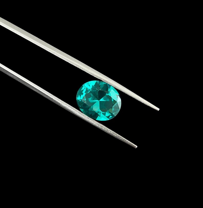 May include: A faceted oval gemstone with a vibrant teal-green color held between the two thin, silver-toned metal tips of a pair of tweezers. The gemstone displays internal light reflections and geometric cuts across its surface.