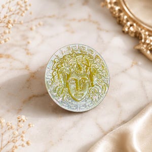 May include: A circular brooch featuring a central relief of a face surrounded by foliage, rendered in shades of olive green and iridescent white. The outer edge is decorated with a geometric Greek key pattern, creating a distinct contrast.