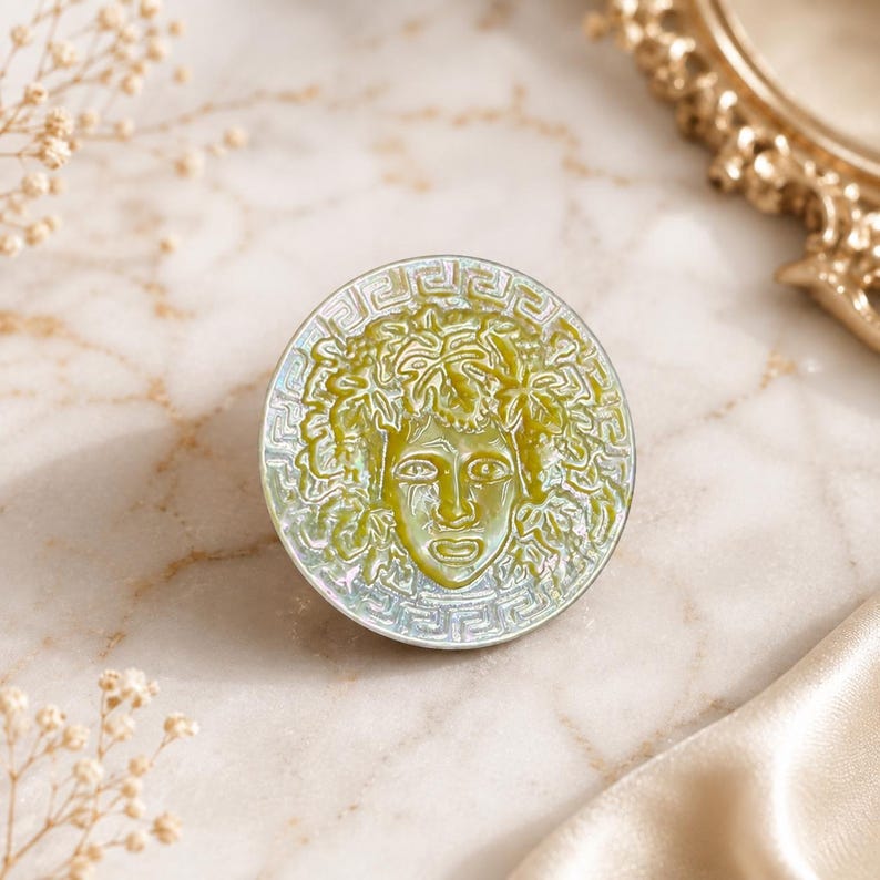 May include: A circular brooch featuring a central relief of a face surrounded by foliage, rendered in shades of olive green and iridescent white. The outer edge is decorated with a geometric Greek key pattern, creating a distinct contrast.